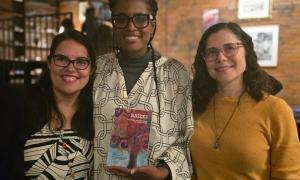 Three women standing. The middle one is holding the poetry in translation book Raizes