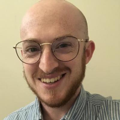 image of smiling white male with glasses and a reddish beard. Raymond is wearing a blue and white striped collared shirt