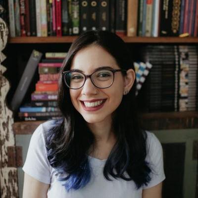 A photo of Laura Vieira, white female wearing glasses and a white t-shirt, dark red lipstick and dark brown hair with blue tips. On the background, a vintage bookcase.
